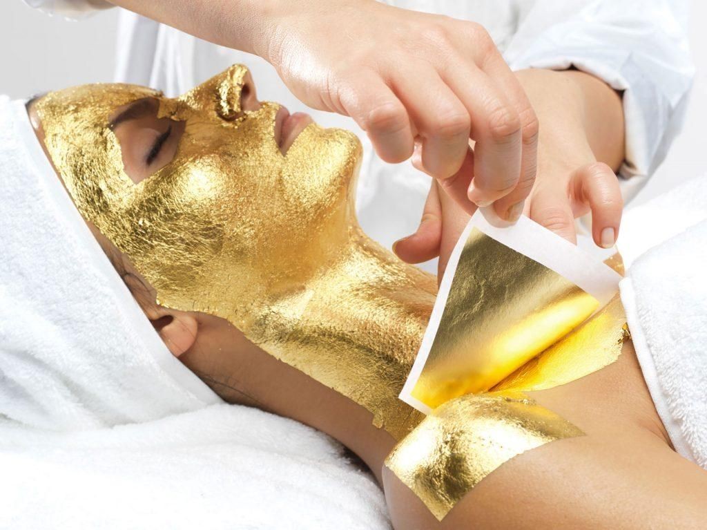 A hand applying gold leaf sheets to a person's skin wrapped in white towels during a spa treatment.